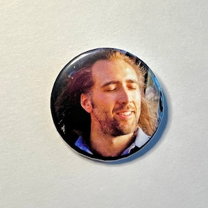 Op de afbeelding: Een ronde knop met een close-up afbeelding van Nicolas Cage met zijn ogen dicht en een glimlach op zijn gezicht. Zijn haar is lang en golvend.