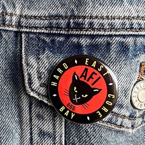 May include: A black and red button with the text "Hard East Bay Core AFI 666" and a black cat with yellow eyes.