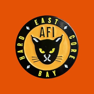 May include: A round, black and yellow pin with the text "EAST AFI BAY HARD CORE" in white lettering. The pin features a black cat with yellow eyes and white whiskers. The background is orange.