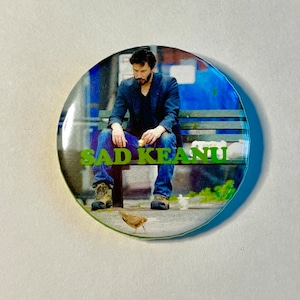 May include: A circular badge with an image of a man sitting on a bench, with the text "SAD KEANU" in green. The man is wearing a dark blazer, blue jeans, and boots. A bird is in the foreground.