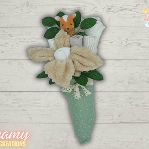 May include: A green and white diaper cake bouquet with a brown deer toy, white and gray washcloths, and a white ribbon. The bouquet is on a white wood background.