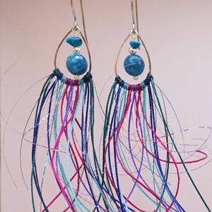 May include: A pair of dangle earrings with blue agate stones and a fringe of colorful threads. The earrings are made with silver wire and have a bohemian style.