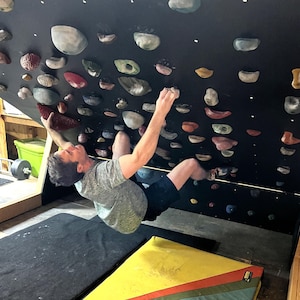 Puede incluir: Una persona escalando en un muro de escalada negro con varios agarres de colores. El muro está inclinado y la persona está alcanzando un agarre. El muro de escalada está en una habitación con una estera negra y una estera amarilla y roja.