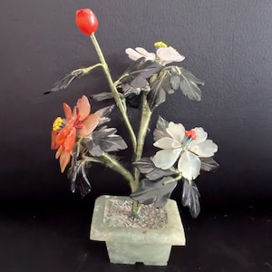 Puede incluir: Un pequeño bonsái de piedra color jade con flores en tonos naranja, blanco y rojo. Las hojas son de color verde oscuro y las flores tienen centros amarillos. El árbol está en una maceta cuadrada, también de piedra color jade.