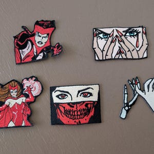 May include: Five embroidered patches featuring comic-style illustrations: a red-clad figure in a headpiece, a face with hands covering eyes, a full-body figure in a red costume, a face with a red skull mask, and a skeletal hand reaching toward a human hand.