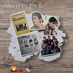 May include: A mystery pack of stickers featuring images from "The Outsiders" film. The pack includes stickers with film stills, a newspaper clipping, and the film title. The stickers are on a wooden surface.
