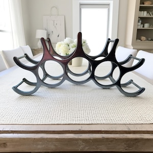 May include: A black and burgundy wine rack with six circular openings. The rack is made of a dark material and has a curved design. It is sitting on a light-colored textured surface.