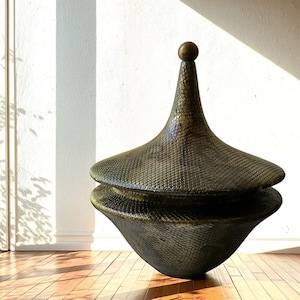 May include: A large, dark bronze-toned decorative vessel with a textured, scale-like pattern across its surface. The object features a wide, rounded base and a matching lid that tapers upward to a point, topped with a small, smooth spherical finial.