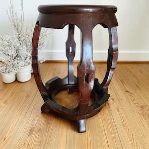 May include: A dark brown wooden stool with a circular top and a curved, barrel-shaped frame. The structure features four vertical legs connected by a circular base and decorative cutouts, showcasing a polished finish and visible wood grain.