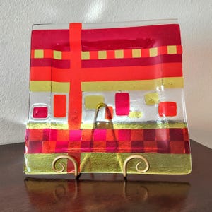 May include: A square glass serving plate with a geometric design in red, orange, and gold. The plate is on a stand.