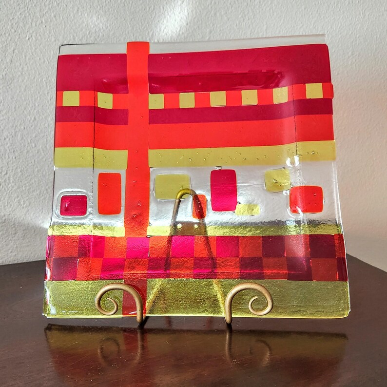 May include: A square glass serving plate with a geometric design in red, orange, and gold. The plate is on a stand.