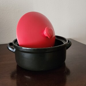 May include: A red, egg-shaped kitchen gadget with a heart-shaped handle is nestled inside a black ceramic pot with two handles. The pot sits on a dark wooden surface, creating a contrast of colors and textures.