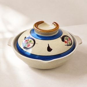 May include: A ceramic Japanese donabe pot with a lid. The pot is beige with a blue stripe and decorative painted accents in red, green, and black. The lid has a brown handle and a blue stripe. The pot has two side handles.