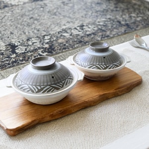 May include: Two ceramic cooking pots with lids. The pots are white with gray lids featuring a geometric design. They sit on a wooden serving board. The background includes a patterned rug and a white tablecloth.