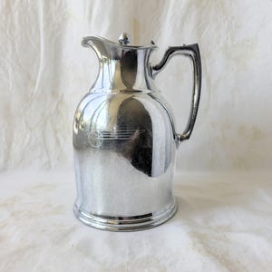 May include: A shiny, silver-colored pitcher with a lid and a curved handle. The pitcher has a rounded body and a spout for pouring. A small emblem is visible on the front. The pitcher is likely made of metal and reflects the surrounding light.
