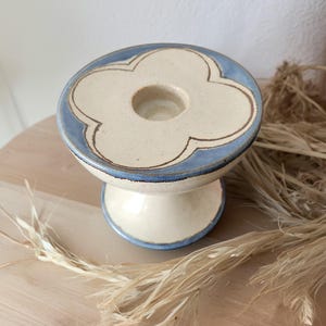 May include: A ceramic candle holder with a cream-colored base and a light blue rim. The top features a four-leaf clover design with a central hole for a candle. The holder is approximately 4 inches tall.