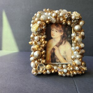 May include: A rectangular picture frame decorated with an assortment of gold-toned beads, white pearls, and clear crystals. The frame holds a portrait of a person with long brown hair, captured in a soft, painterly style with muted tones.
