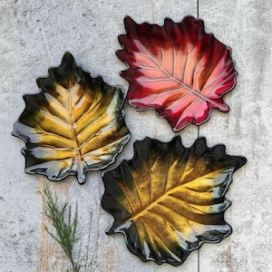 May include: Three decorative leaf-shaped dishes in shades of red, gold, and black. The dishes have a glossy finish and detailed veining, resembling autumn foliage. The arrangement is on a light-colored, textured surface.