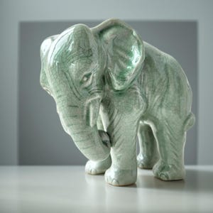 May include: A ceramic elephant figurine featuring a light mint green glaze with a prominent dark crackle pattern throughout the surface. The elephant is depicted in a standing position with its trunk curved downward toward its front legs.
