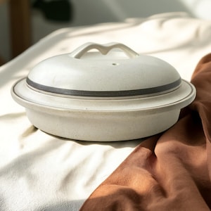 May include: A speckled, off-white ceramic casserole dish with a matching lid. The lid has a handle and a dark gray band around the edge. The dish sits on a white and brown fabric.