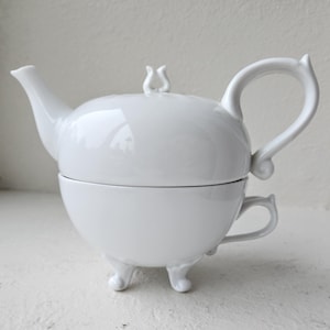 May include: A white ceramic teapot and teacup set. The teapot sits atop the teacup, with a curved handle and a small, decorative finial on the lid. The teacup has a small handle and three small feet.