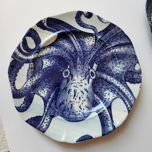 May include: A round ceramic plate with an irregular, wavy edge featuring a detailed dark blue ink-style illustration of an octopus. The design covers the entire surface, showcasing the creature's head, eyes, and multiple tentacles in deep blue.