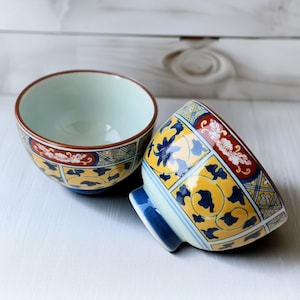 May include: Two ceramic bowls with a yellow, blue, and red floral pattern. The bowls have a light blue interior and a brown rim. One bowl is upright, and the other is tilted on its side.