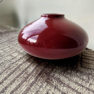 May include: A glossy, maroon-colored ceramic vase with a rounded body and a small opening. The vase sits on a textured, neutral-toned surface, reflecting light. The design is simple and elegant.