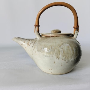 May include: A ceramic teapot with a light beige glaze and brown speckles. It features a curved spout, a small lid, and a handle made of woven light brown material. The teapot is designed for brewing tea.