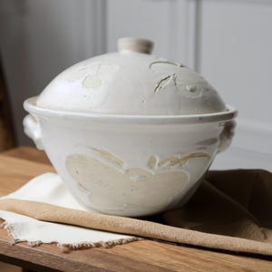 May include: A cream-colored ceramic serving bowl with a lid. The bowl features embossed apple designs and handles. The lid has a small knob. The bowl is sitting on a wooden surface with a beige cloth and a wooden spoon.