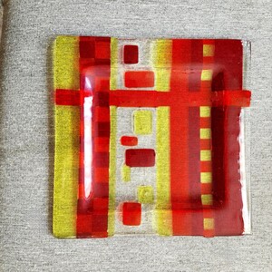 May include: A square glass plate with a geometric design in red and yellow. The plate has a clear glass centre with red and yellow squares and rectangles layered on top.