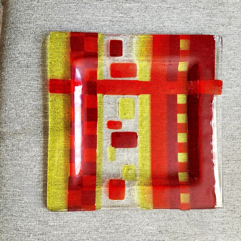 May include: A square glass plate with a geometric design in red and yellow. The plate has a clear glass centre with red and yellow squares and rectangles layered on top.