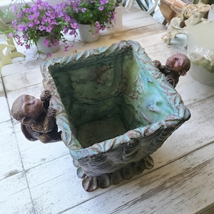 May include: A square planter with a weathered teal and brown finish, featuring two sculpted monkey figures clinging to opposite sides of the rim. The exterior has a textured, leaf-like pattern, and the base is supported by four rounded feet.