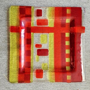 May include: A square glass plate with a geometric design in red and yellow. The plate has a clear glass border and a textured surface.