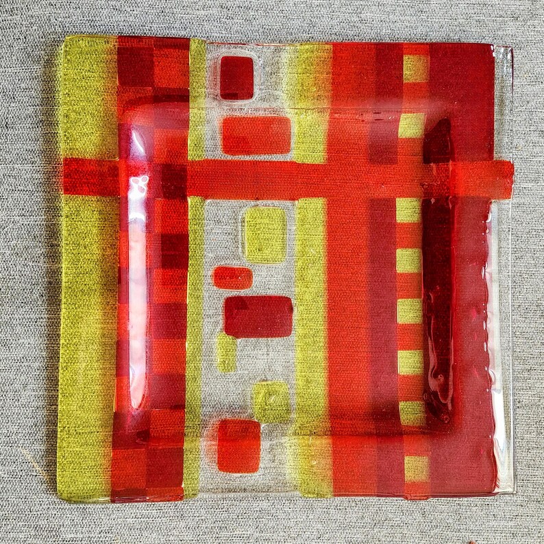 May include: A square glass plate with a geometric design in red and yellow. The plate has a clear glass border and a textured surface.