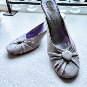 May include: A pair of light gray suede heeled shoes with a bow detail on the toe. The shoes have a purple lining and the brand name "CONNI" is printed on the heel.