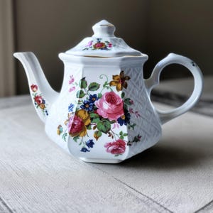 May include: White porcelain teapot with a floral design featuring pink roses, blue and yellow flowers, and green leaves. The teapot has a geometric shape with a lid and a curved handle. The spout and lid are adorned with additional floral accents.