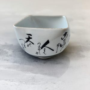 May include: A square-shaped ceramic bowl with a white glazed finish and black hand-painted Japanese calligraphy characters wrapping around the exterior sides. The bowl features a wide, open top and a tapered base with a smooth, clean aesthetic.