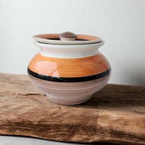 May include: A ceramic jar with a lid, featuring horizontal bands of orange, black, and brown. The jar has a white rim and a small, brown knob on the lid. The jar is sitting on a wooden surface.