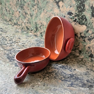 May include: Two terracotta-colored ceramic bowls. One bowl has a handle, and the other has a small ear-shaped handle. The interior of the bowls is a lighter shade of orange. The bowls are likely for serving or cooking.
