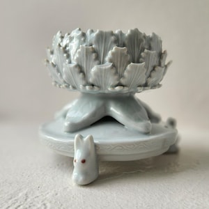 May include: A light blue ceramic decorative bowl with a detailed, textured design. The bowl sits atop a pedestal with a small, white rabbit figurine with red eyes attached to the base. The bowl has a floral design.