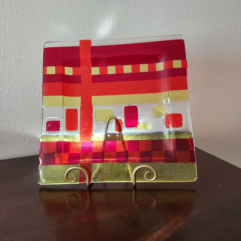 May include: A square glass serving dish with a red, gold and white geometric design. The dish is on a gold metal stand.