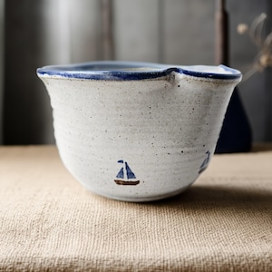 May include: A speckled white ceramic bowl with a blue rim and small sailboat illustrations. The bowl has a unique, wavy top edge and a rustic, handmade appearance. The sailboats are blue with brown hulls.