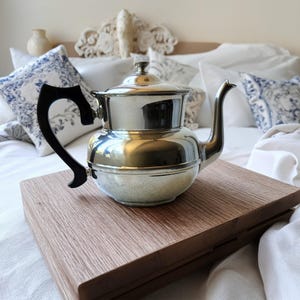 May include: A polished silver-tone metal teapot featuring a rounded body, a curved spout, and a black handle. The teapot has a matching lid with a small knob at the top. The reflective surface shows light highlights, and the design is simple and functional.