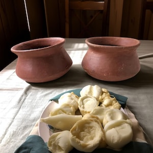 May include: Two terracotta-colored clay pots with wide rims sit on a table. Below, a plate holds various white, soft cheese pieces. The scene is lit by natural light, suggesting a rustic, home-cooked meal.