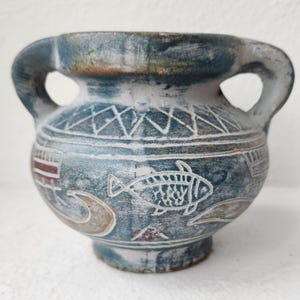 Puede incluir: Un pequeño jarrón de cerámica azul verdoso con dos asas. El jarrón presenta diseños blancos grabados, incluyendo un pez, patrones geométricos y otras formas abstractas. El jarrón tiene una apariencia desgastada.