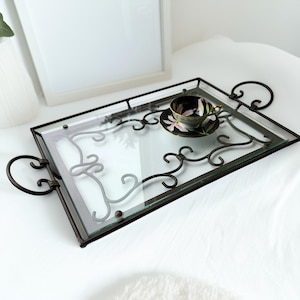 May include: A rectangular serving tray with a glass top and dark metal frame. The tray features decorative scrollwork and curved handles. A teacup and saucer with a floral design rest on the tray.