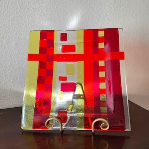 May include: A square glass plate with a geometric design featuring red, yellow and white stripes and squares. The plate is on a gold metal stand.