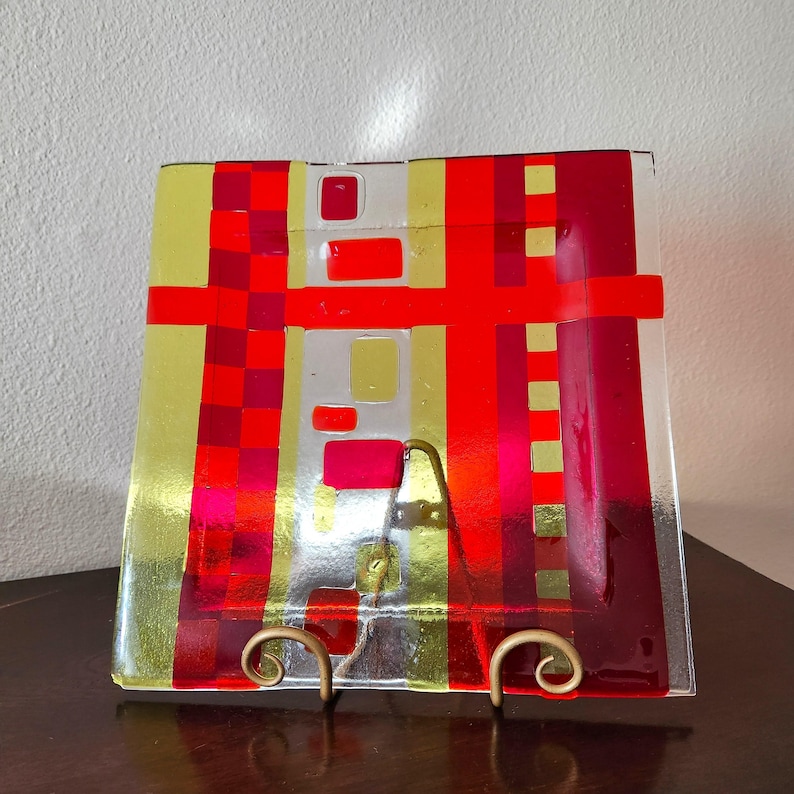 May include: A square glass plate with a geometric design featuring red, yellow and white stripes and squares. The plate is on a gold metal stand.