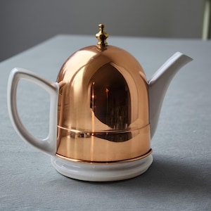 May include: A copper-colored teapot with a white handle, spout, and base. The lid is topped with a small gold finial. The teapot has a reflective surface, and the design is a mix of modern and classic styles.